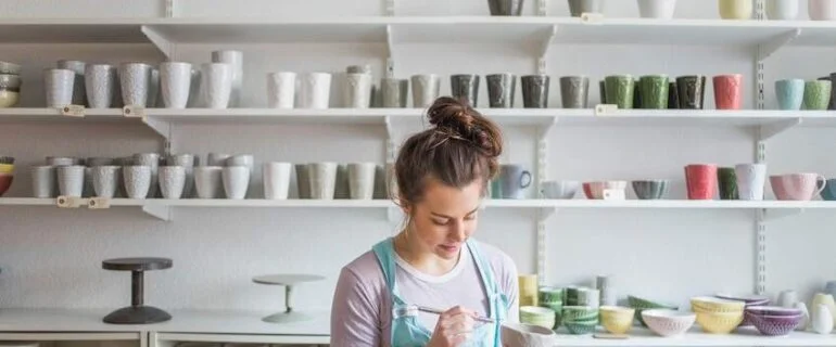 Young female potter using hand tool on vase while standing against shelves at store