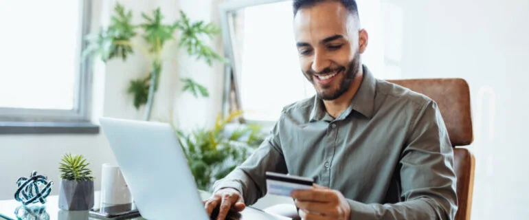 A businessman in his office smiles as he consults his credit card and looks at his laptop