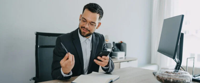 A professional man in a suit seated at a desk, engaged with his smartphone and holding a credit card