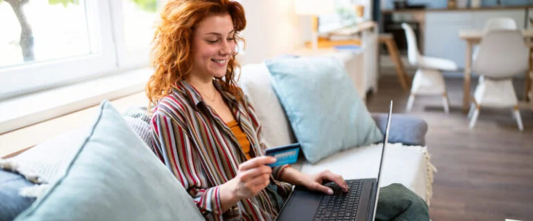 A young woman sitting on the sofa holding a credit card in one hand as she balances her laptop on her knee
