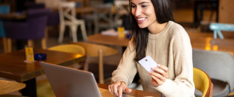 A young woman smiles holding a credit card while using laptop and sitting in a cafe.
