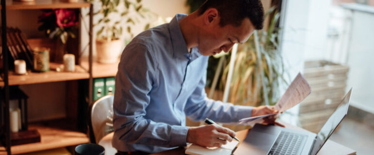 A man with a calculator looks over a bill with his laptop open on the desk before him