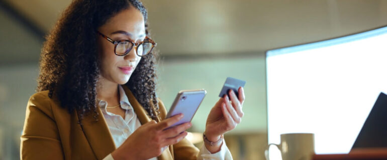 A woman sitting at her desk with a laptop open in front of her and a mug on the desk holds a credit card in one hand and her mobile phone in the other
